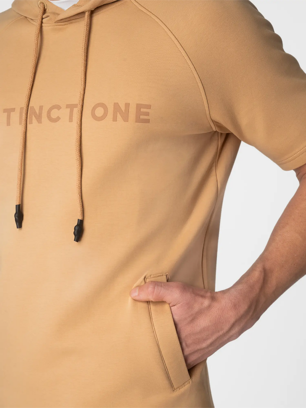 A close-up of a tan pullover hoodie worn by a model, showing the chest with the brand name 'DISTINCTONE' in a matching tan print, black drawstrings, and a side pocket with the model's hand inside.