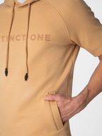 A close-up of a tan pullover hoodie worn by a model, showing the chest with the brand name 'DISTINCTONE' in a matching tan print, black drawstrings, and a side pocket with the model's hand inside.
