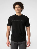 A man wearing a black short-sleeve t-shirt with 'DISTINCT ONE' printed in a subtle dark gray font across the chest, paired with dark gray pants, against a light gray studio background.