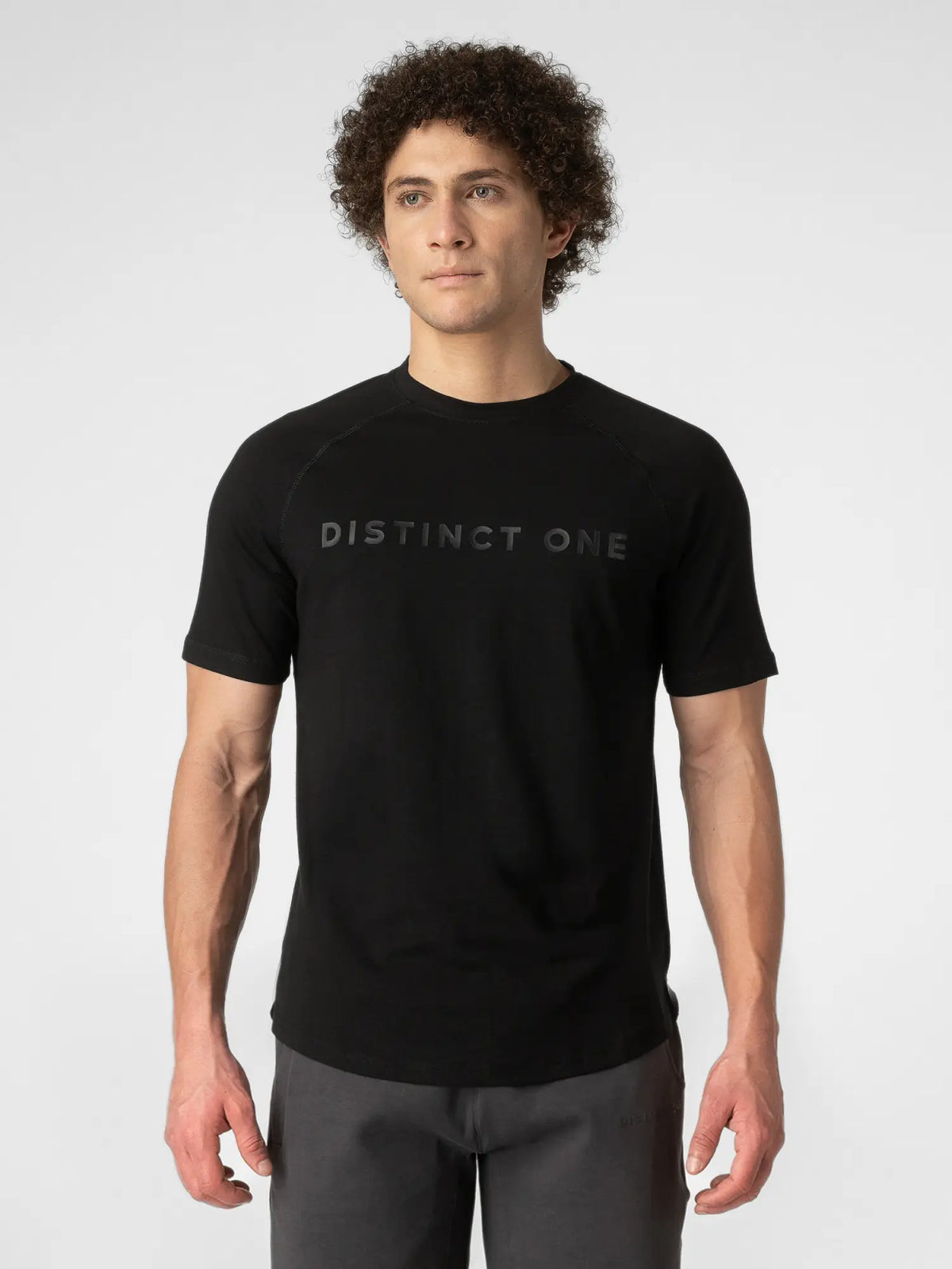 A man wearing a black short-sleeve t-shirt with 'DISTINCT ONE' printed in a subtle dark gray font across the chest, paired with dark gray pants, against a light gray studio background.