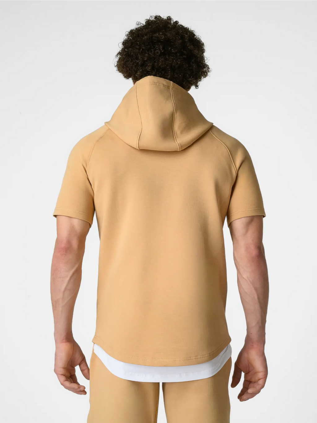 A man modeling a matching tan short-sleeve hoodie and shorts set with white crew socks and gray sneakers, standing against a light gray studio background.