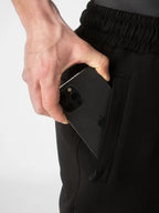 A close-up of a person's hand pulling a black smartphone out of a side pocket of black athletic pants, shown against a clean white background.