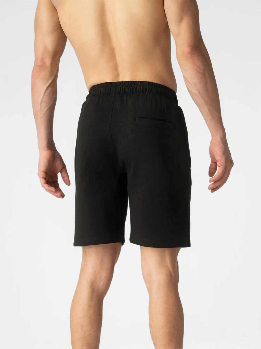 Rear view of a man wearing black athletic shorts against a light gray studio background.