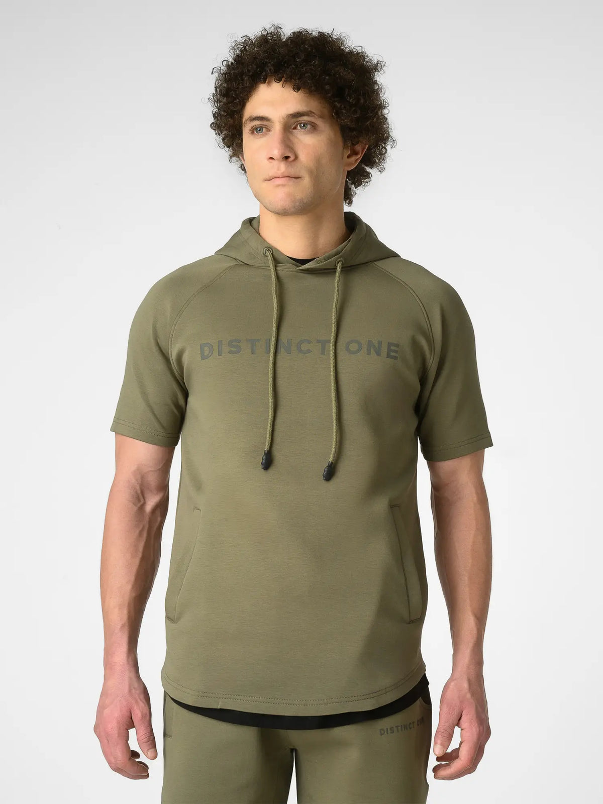 A man wearing an olive green short-sleeve hoodie with 'DISTINCTONE' printed across the chest, paired with matching olive green pants, photographed against a light gray studio background.