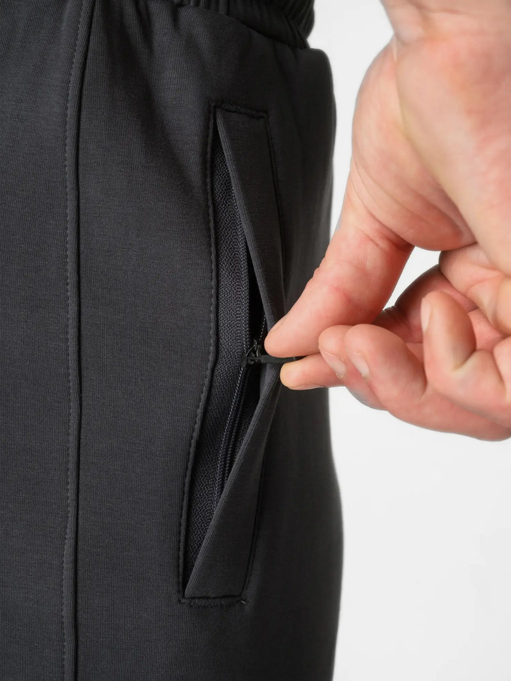 Close-up of a person's hand opening a zippered side pocket on black athletic pants against a light gray background.