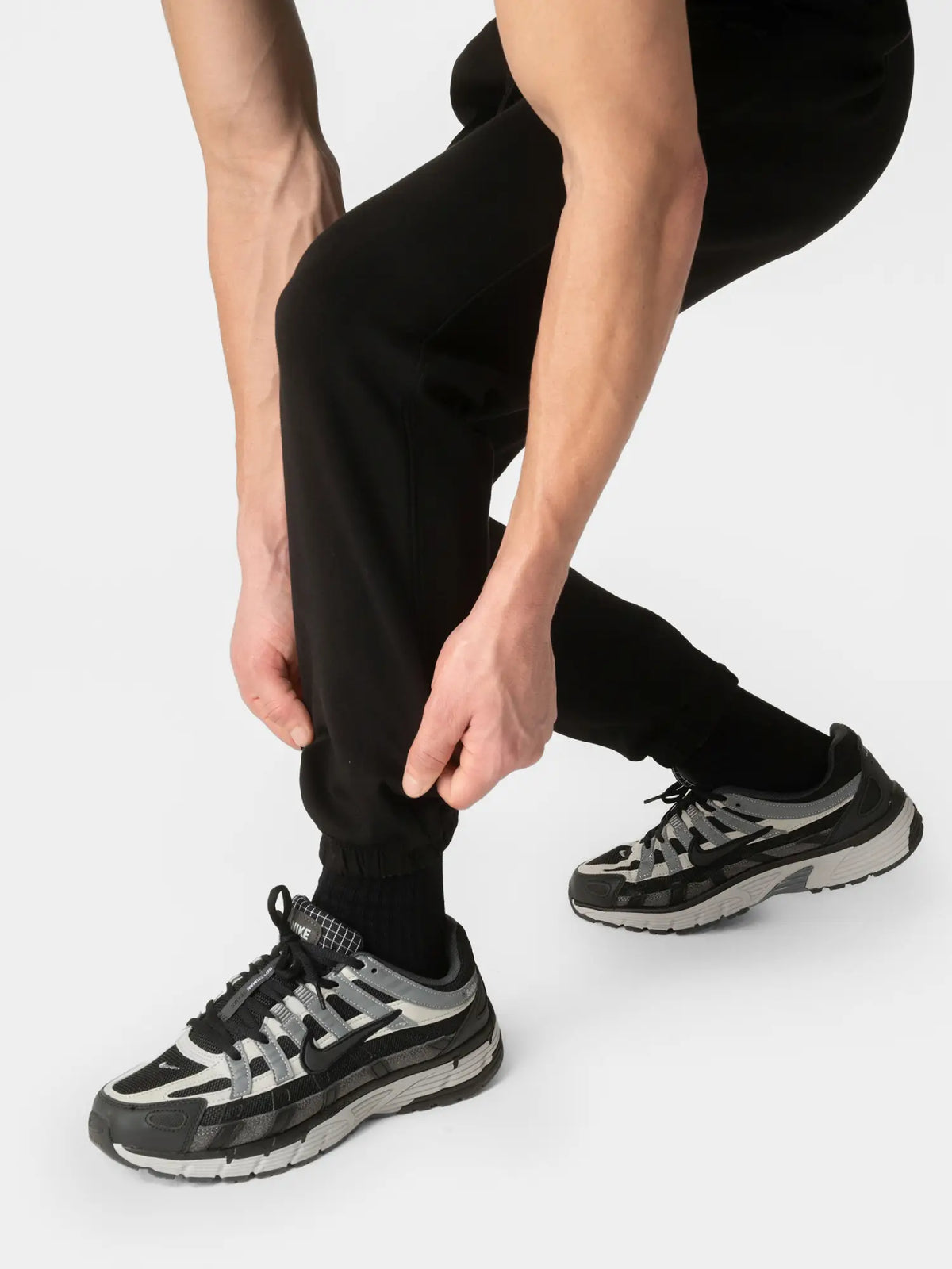 A close-up of a person wearing black jogger pants and black athletic sneakers, adjusting the cuff of the jogger with both hands against a light gray studio background.