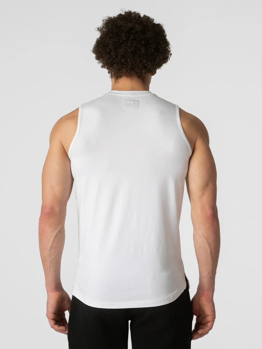 A man viewed from behind wearing a white sleeveless athletic tank top and black pants, against a light gray studio background.