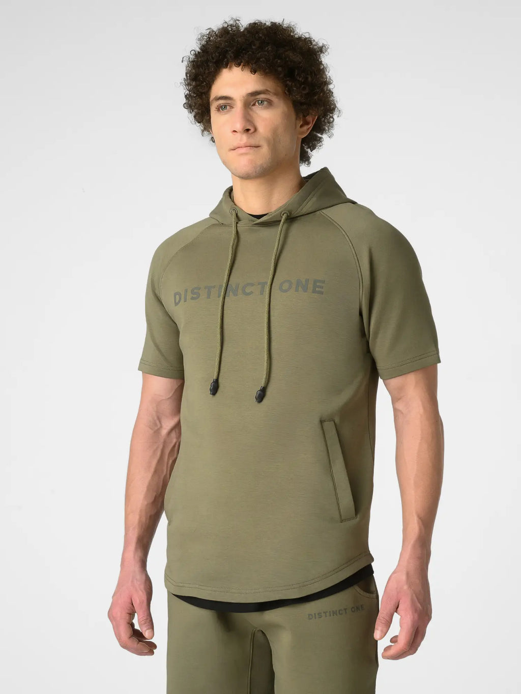A man wearing an olive green short-sleeve hoodie with 'DISTINCTONE' printed in gray across the chest, paired with matching olive green pants, photographed against a light gray studio background.