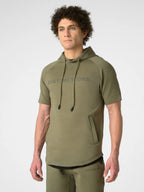 A man wearing an olive green short-sleeve hoodie with 'DISTINCTONE' printed in gray across the chest, paired with matching olive green pants, photographed against a light gray studio background.