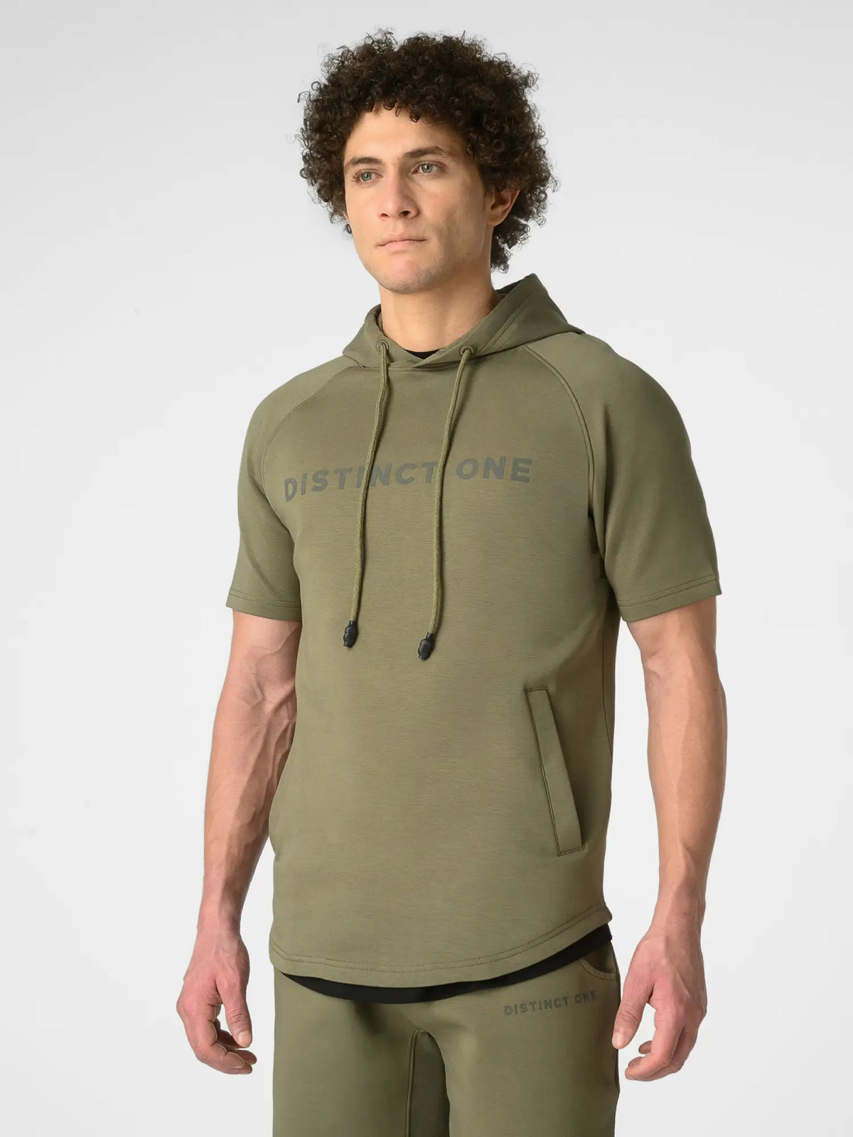 A man wearing an olive green short-sleeve hoodie with 'DISTINCTONE' printed in gray across the chest, paired with matching olive green pants, photographed against a light gray studio background.