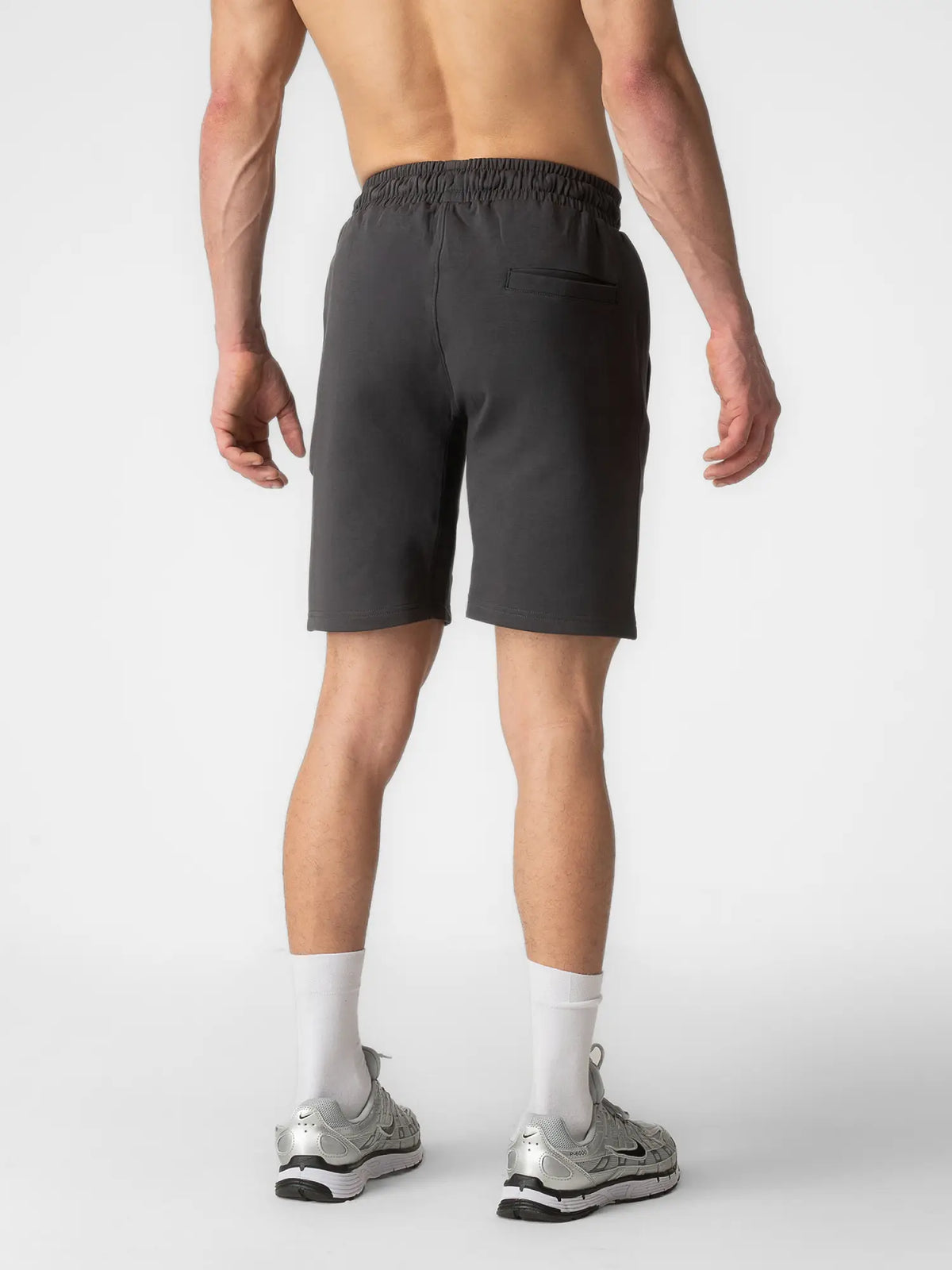 A man shown from the back wearing dark gray athletic shorts, white crew socks, and silver running shoes against a light gray studio background.
