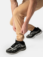 A close-up of a person wearing beige jogger sweatpants and black and gray athletic sneakers, crouching down to adjust the cuffs of the joggers against a light gray studio background.