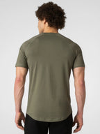 A man viewed from behind wearing an olive green short-sleeve athletic t-shirt and black pants, set against a plain light gray studio background.