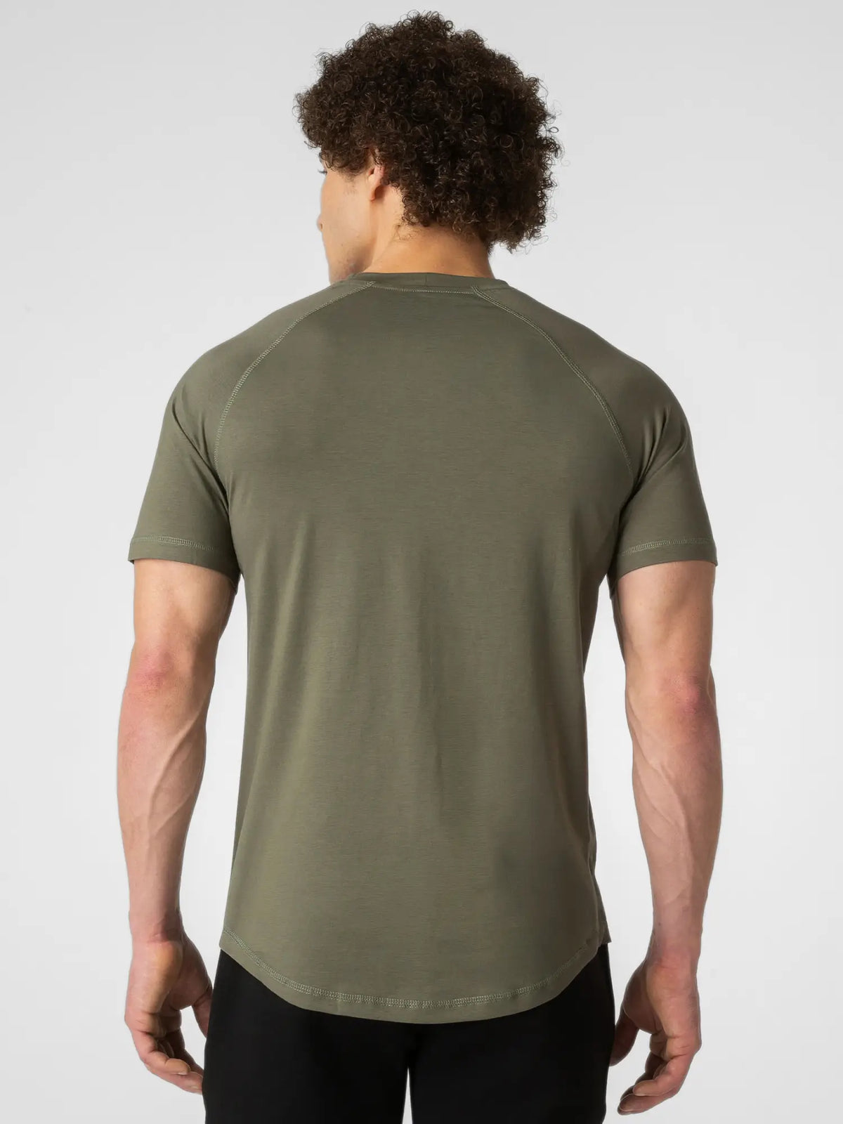 A man viewed from behind wearing an olive green short-sleeve athletic t-shirt and black pants, set against a plain light gray studio background.