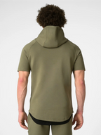 A man viewed from behind wearing a short-sleeve olive green hooded athletic shirt and matching green pants against a light gray studio background.