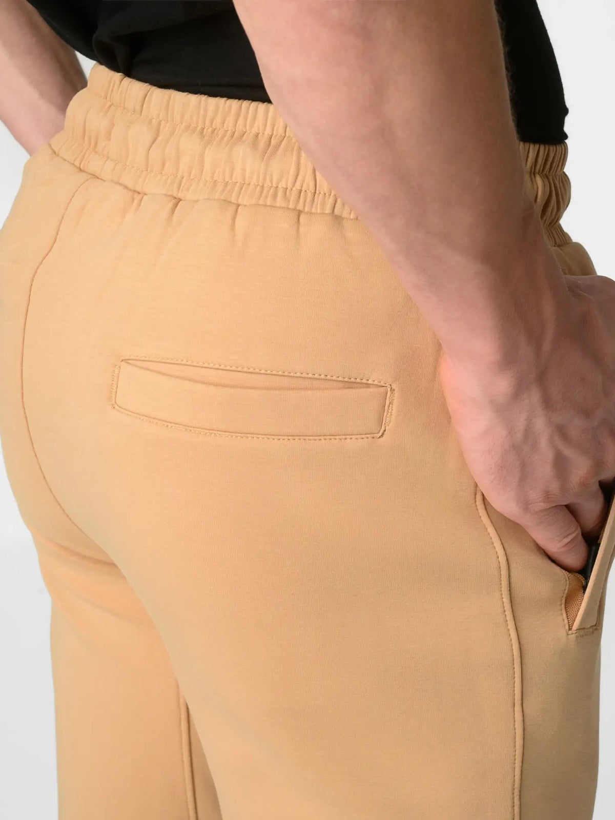 Close-up of a person wearing tan or camel-colored drawstring pants with a side pocket, paired with a black top, against a light gray studio background.