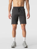A man wearing dark gray athletic shorts, white socks, and gray sneakers, shown from the waist down against a light gray studio background.