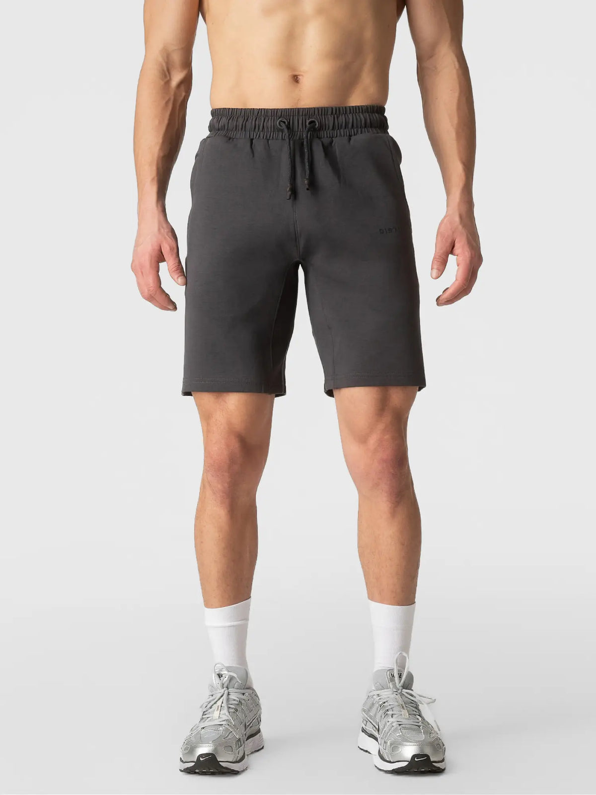 A man wearing dark gray athletic shorts, white socks, and gray sneakers, shown from the waist down against a light gray studio background.